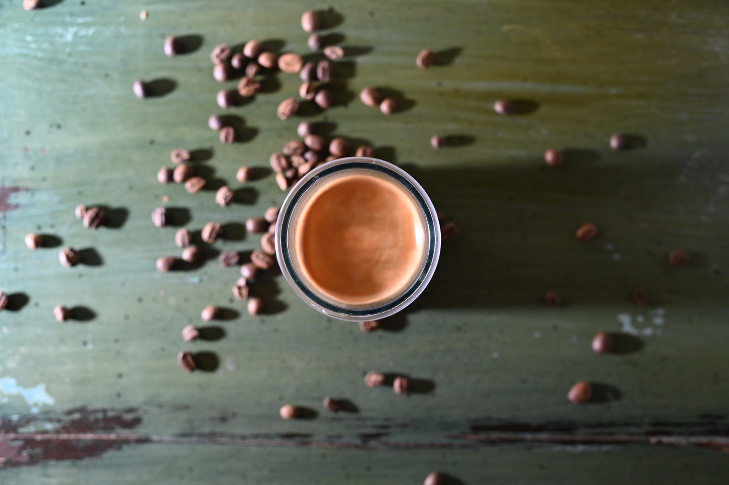Aerial view of espresso cup on a green rustic table with scattered coffee beans.