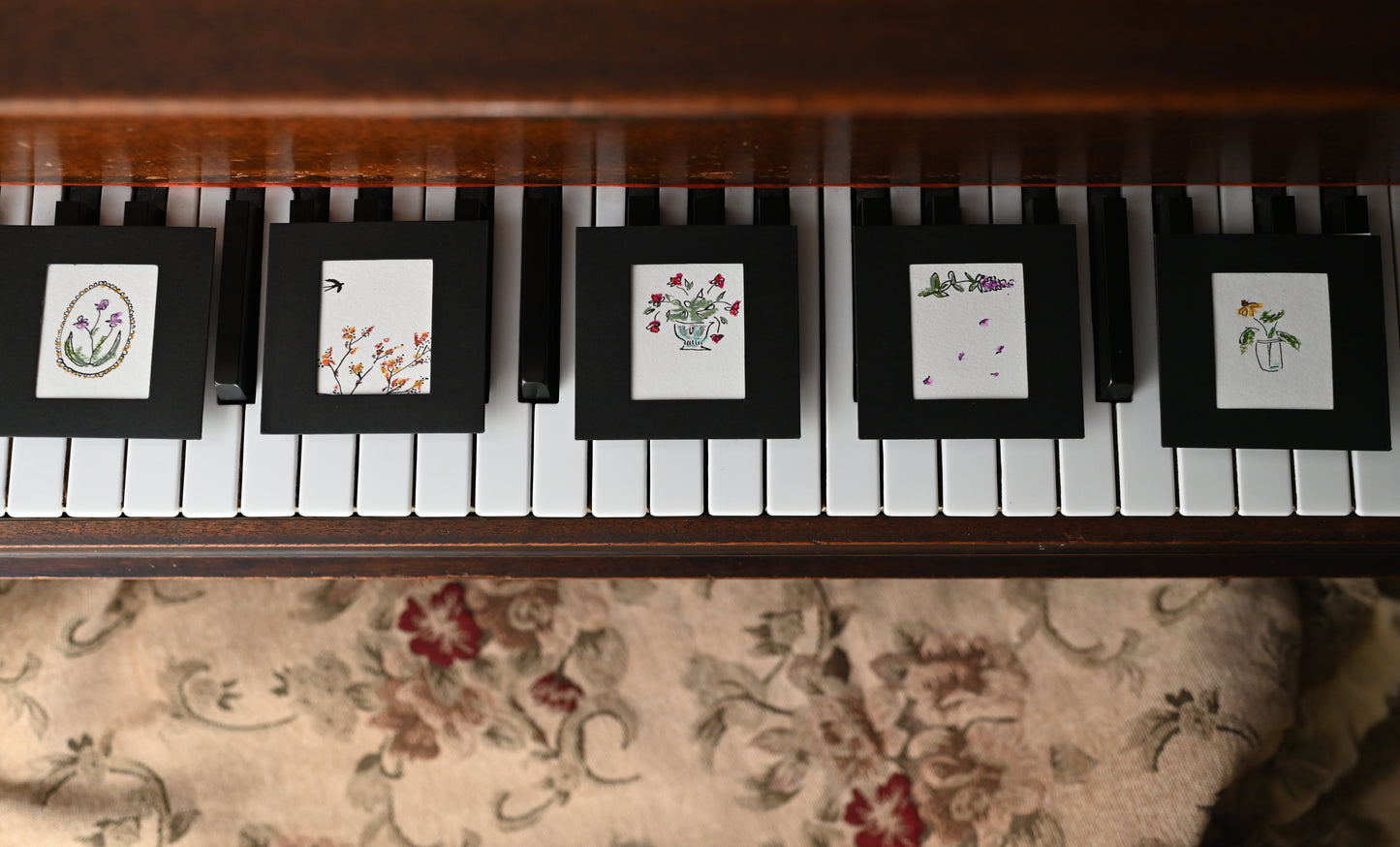 Set of five floral watercolor art cards displayed on piano keys, featuring vibrant flowers in black frames.
